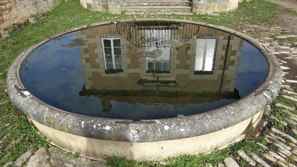 Bassin circulaire en pierre rempli d’eau, dans lequel se reflète la façade d’une maison.