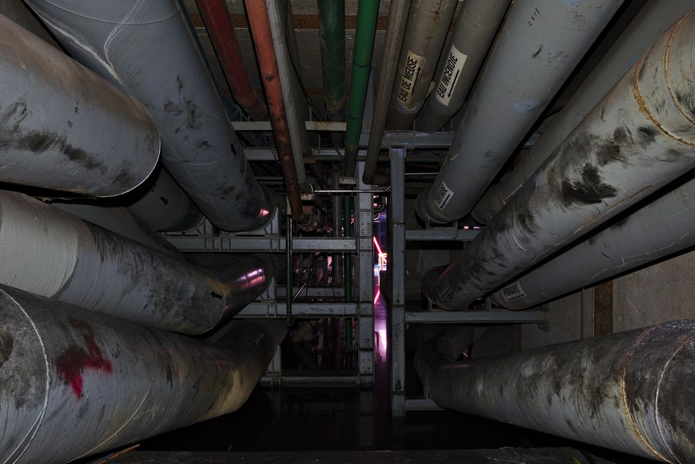 Galerie technique d’une installation de traitement d’eau, conduites calorifugées étiquetées « eau de Meuse » et « eau incendie ».